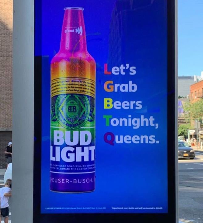 A bud light advert on a bus stop for pride. It has a rainbow beer bottle with the words Let's Grab Beers Tonight, Queens